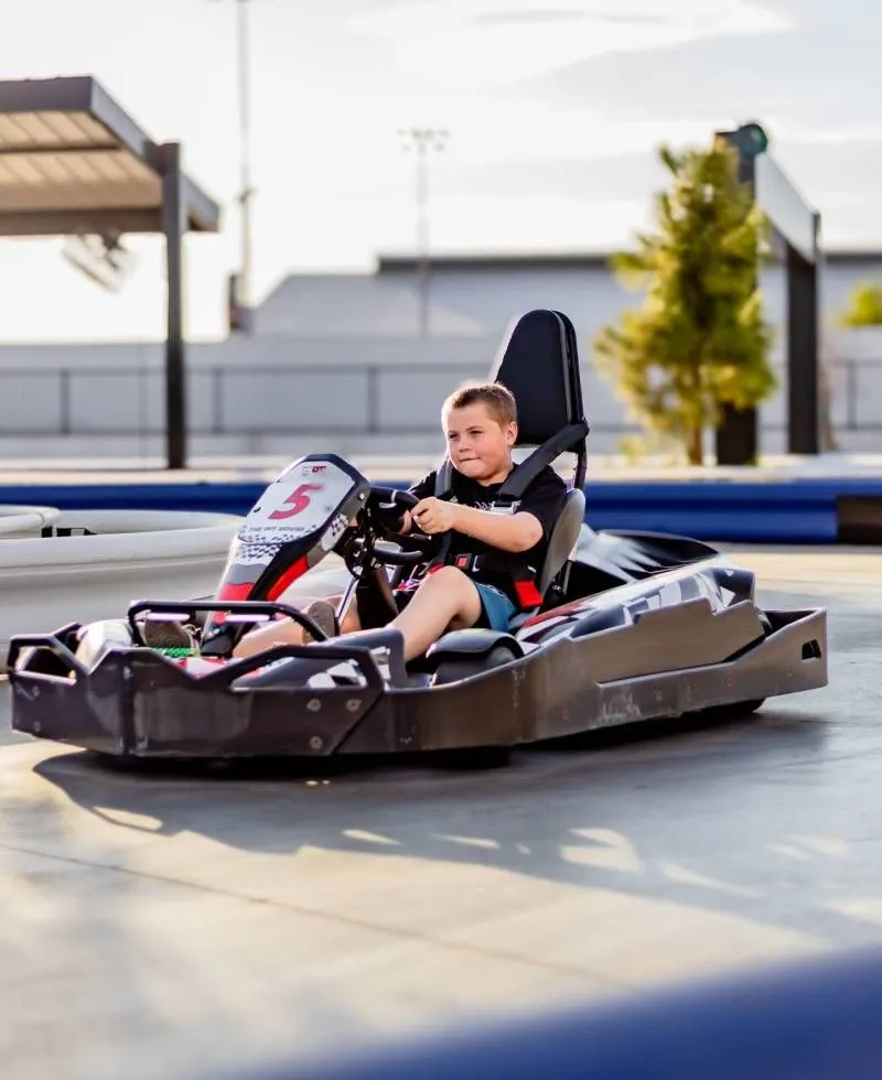 Child driving a black go-kart on an outdoor track, with a blurred background and trees.