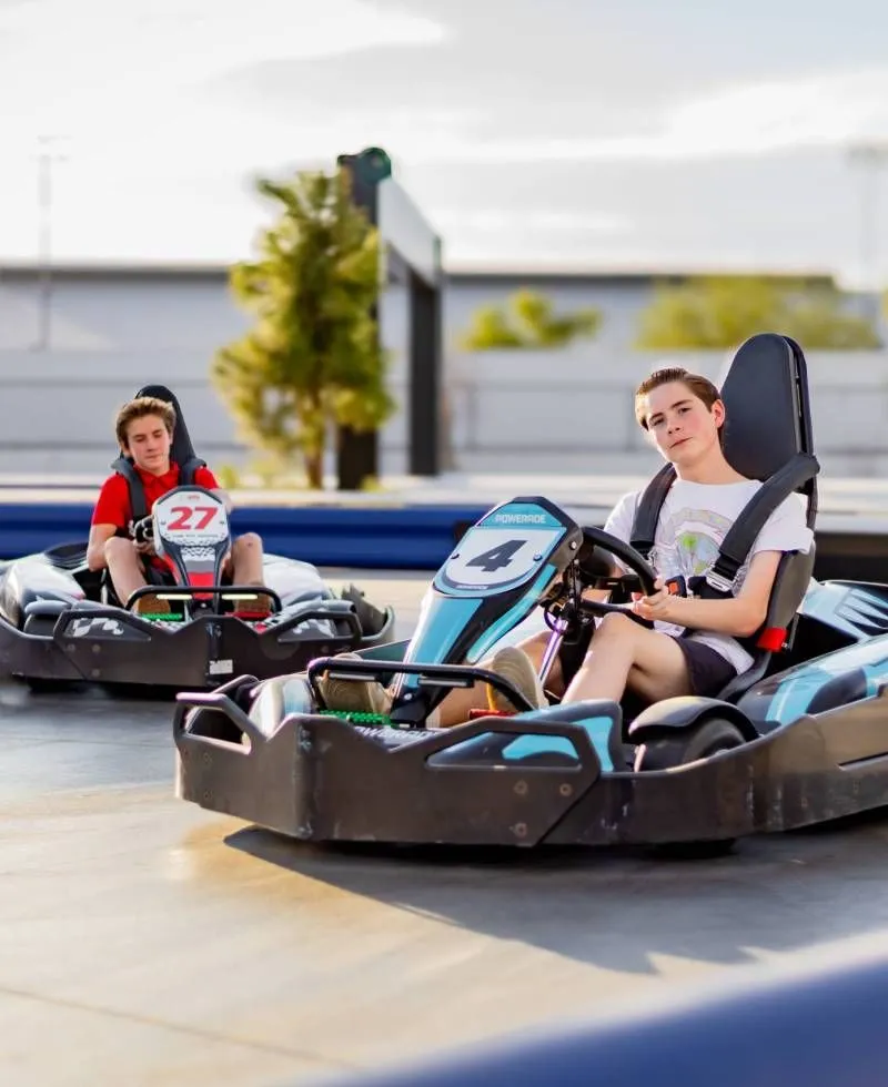 Two people driving black go-karts on an outdoor track, one in the foreground wearing a white shirt.