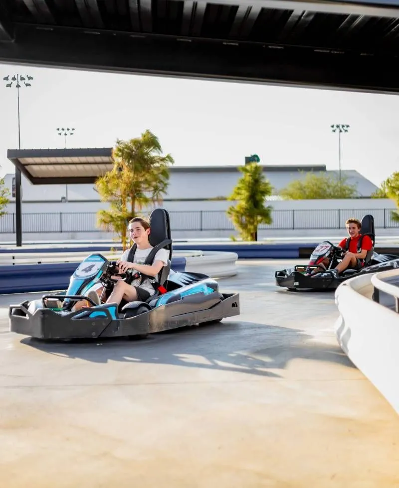 Two people driving electric go-karts on an outdoor track, one blue kart in front and one red kart behind