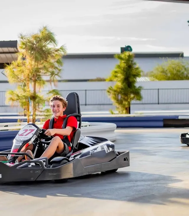 Child driving a red go-kart on an outdoor track, wearing a black helmet and smiling.