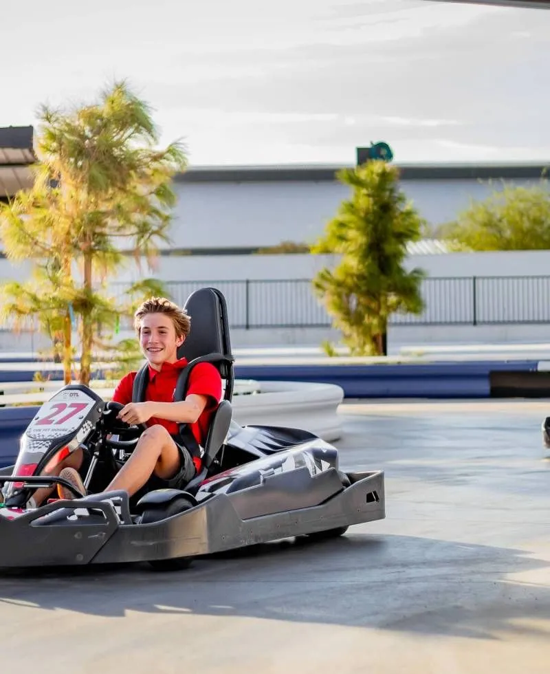 Person in a red shirt driving a go-kart on a track, smiling with trees and a building in the background.