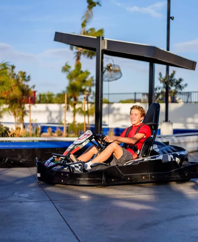 Person seated in a black go-kart under a canopy at an outdoor track, wearing a red shirt.