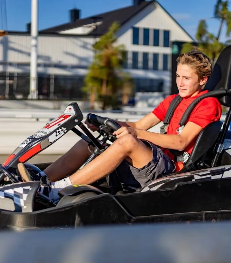 Young person driving a go-kart on a track, wearing a red shirt and helmet-less, with buildings in the background