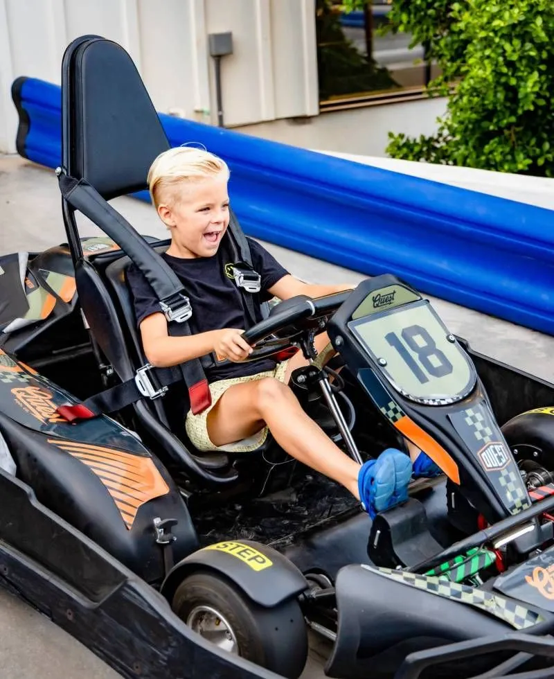 Child smiling in a black go-kart with orange accents, racing on a track.