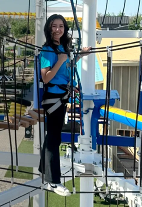 Person in a harness smiles on a high ropes course, with blue and yellow equipment behind.