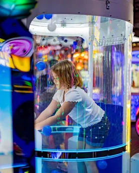 Child playing in a colorful arcade game booth with glowing lights and a transparent enclosure