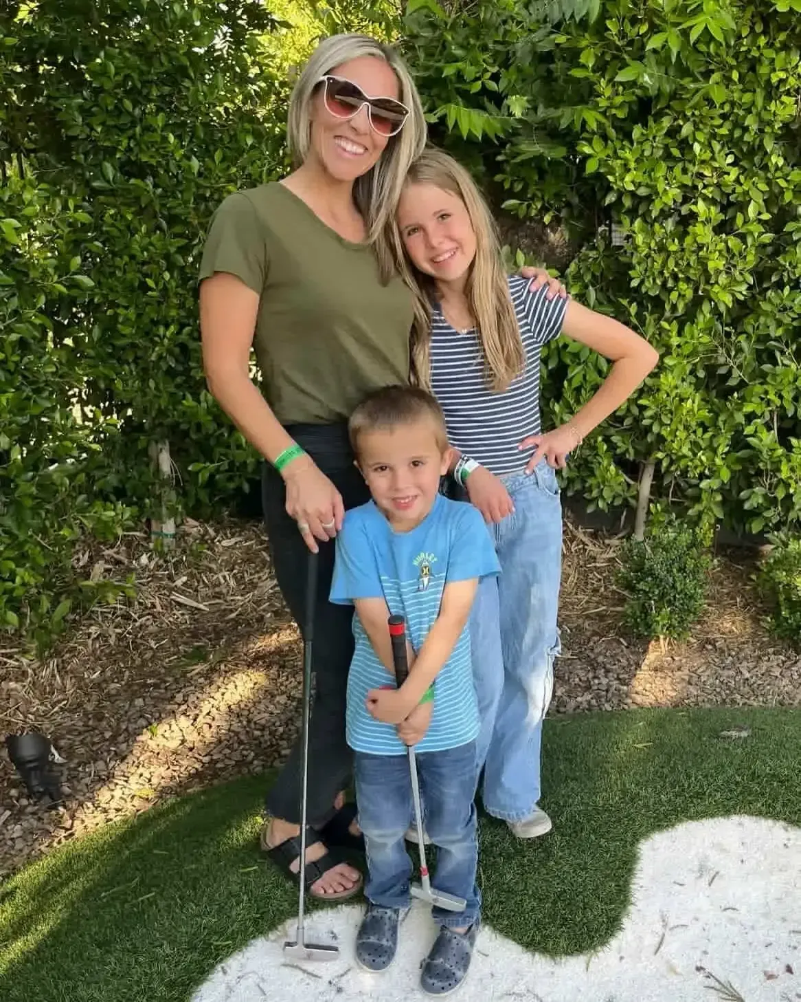 Woman and two children posing outdoors in front of a green hedge, smiling by a white-painted curb.