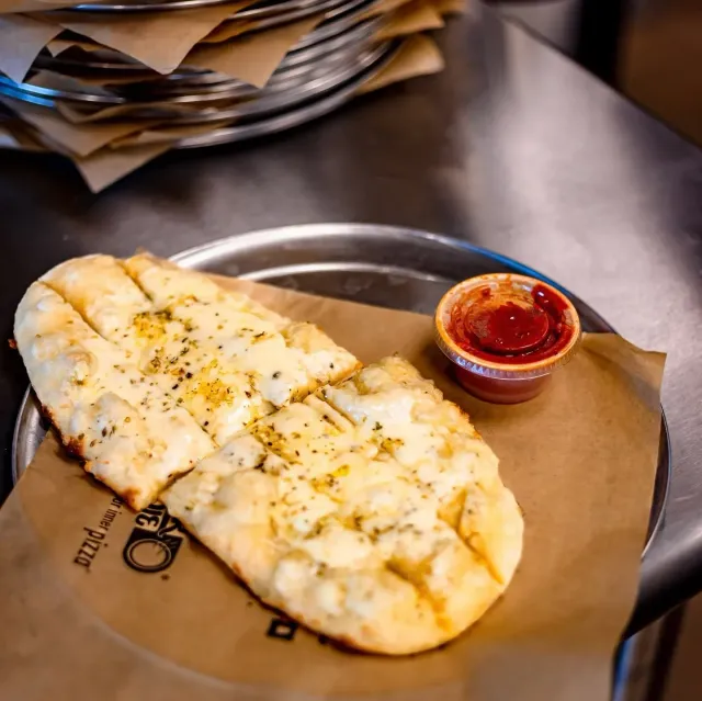Cheesy flatbread on brown paper with a small cup of red dipping sauce on a metal table
